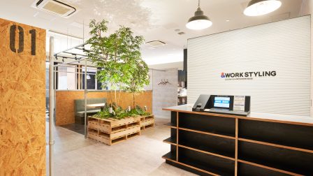 武蔵小杉駅東口から徒歩1分、ららテラス武蔵小杉4階に位置するワークスタイリングららテラス武蔵小杉店のエントランス。木目の受付カウンターにはロゴとチェックイン用の端末が設けられ、中央にはシンボルツリーの観葉植物が配置されている。奥にはOSB合板で仕切られた半個室のソファブースが見え、コワーキングスペースとして落ち着いて利用できる空間となっている