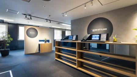 ワークスタイリング四谷のエントランス・受付エリアの画像。木目調カウンターと受付端 末、WORK STYLINGのサインが見える落ち着いた内装で、飯田橋駅周辺のワークスタイリ ング拠点とあわせて利用を検討しやすい周辺拠点